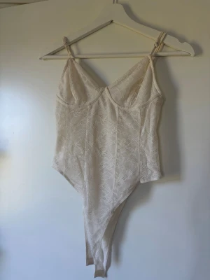 Vit spetsbody från Stradivarius - Säljer en vit body från Stradivarius i spets med tunna axelband och v-ringad urringning. Bodyn har ett fint blommigt spetsmönster och ger en feminin och trendig look. Perfekt att styla med jeans eller kjol för en snygg outfit.