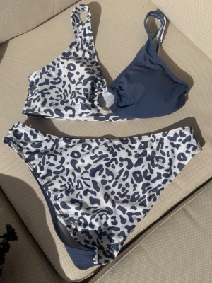Leopardmönstrad bikini i blått och vitt - Snygg bikini med leopardmönster i blått och vitt. Överdelen har en cool asymmetrisk design med en ringdetalj och breda axelband. Underdelen är högmidjad och matchar toppen med samma mönster och färg. Perfekt för stranden eller poolens!