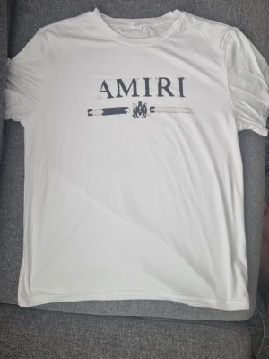 Vit AMIRI t-shirt med tryck - Säljer en vit t-shirt från AMIRI med svart tryck framtill. T-shirten har klassisk passform, rund hals och korta ärmar. Materialet känns mjukt och skönt, perfekt till vardags. Stort AMIRI-tryck och grafisk detalj på bröstet.