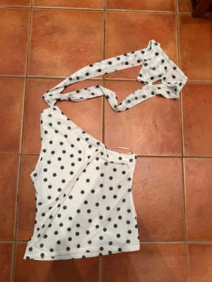 Prickig oneshoulder top - Snygg vit oneshoulder topp med svarta prickar och långt knytband som kan stylas på olika sätt. Toppen är ärmlös och har en cool asymmetrisk design som sticker ut. Perfekt för dig som vill ha något unikt och trendigt i garderoben.