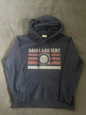 Mörkblå Saint Laurent hoodie - Snygg mörkblå hoodie från Saint Laurent med tryck framtill i rött och vitt samt logga i mitten. Klassisk känguruficka och dragsko i huvan. Perfekt för dig som gillar streetwear och vill ha något stilrent men ändå med attityd.