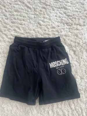 Svarta shorts från Moschino Milano - Svarta shorts från Moschino Milano med vit logotyp och kattmotiv på framsidan. De har elastisk midja och är tillverkade i mjuk bomull, vilket gör dem riktigt bekväma. Perfekta för en avslappnad och cool look.