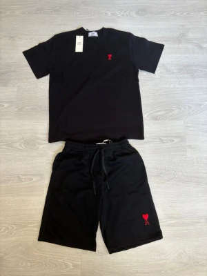 Svart set från Ami Paris med röd logga - Snyggt svart set från Ami Paris med t-shirt och shorts. Båda plaggen har Ami Paris ikoniska röda hjärta med A broderat på bröstet och benet. T-shirten har rund hals och korta ärmar, shortsen har dragsko i midjan. Perfekt för en avslappnad och trendig look.