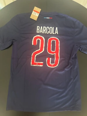 PSG matchtröja Barcola #29 Nike - Mörkblå PSG matchtröja från Nike med Barcola och nummer 29 på ryggen. Framsidan har PSG-logga, Qatar Airways sponsortryck och en röd vertikal rand med vita detaljer. Tillverkad i lätt och ventilerande funktionsmaterial, perfekt för fotboll.