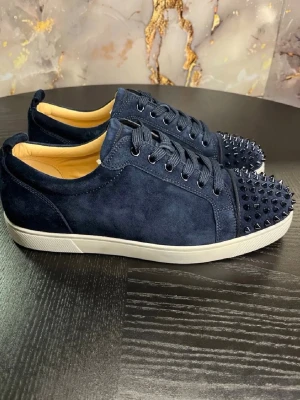 Mörkblå sneakers med nitar - Säljer ett par mörkblå sneakers från Christian Louboutin i mocka med coola svarta nitar på tån. Skorna har vita sulor, matchande mörkblå skosnören och en snygg, stilren design som sticker ut. Perfekta för dig som vill ha något unikt och edgy i garderoben. Dustbag följs med-ingen box.