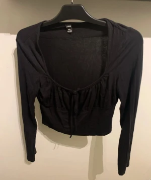 Svart croppad topp med knyt - Trendig svart croppad topp från SHEIN med långa ärmar och djup urringning. Toppen har en söt knytning framtill och lätt rynkade detaljer vid bysten. Perfekt att styla med höga jeans eller kjol.