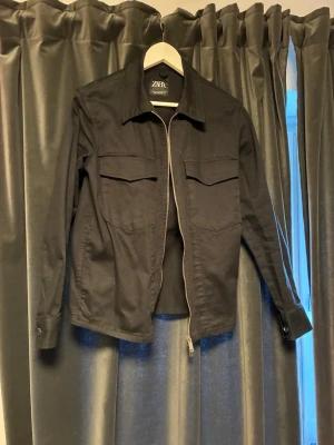 Svart overshirt från Zara - Snygg svart overshirt från Zara med raka linjer och två stora bröstfickor. Jackan har dragkedja framtill, klassisk krage och långa ärmar med knapp vid ärmslut. Perfekt att slänga över en hoodie eller t-shirt för en clean look.