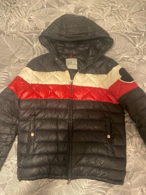 Svart Moncler dunjacka med huva - Snygg svart dunjacka från Moncler med huva och dragkedja. Jackan har breda ränder i vitt och rött över bröstet och armarna samt två fickor med guldfärgade knappar. Klassisk Moncler-logga på ärmen. Perfekt för kalla vinterdagar.