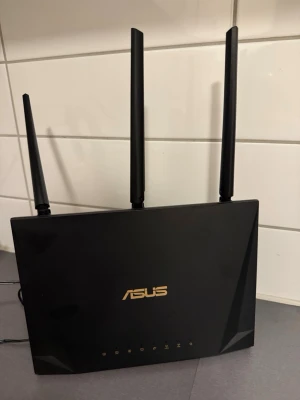 Svart ASUS router med tre antenner - Säljer en svart ASUS router med tre höga antenner och guldfärgad logga på framsidan. Routern har en stilren, rektangulär design i plast och flera indikatorlampor på framsidan. Perfekt för stabilt wifi hemma eller på kontoret.
