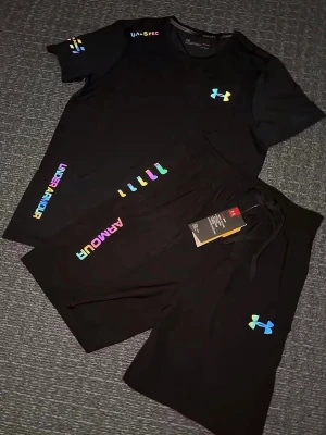 Svart träningsset från Under Armour - Säljer en svart träningsset från Under Armour med färgglada loggor och textdetaljer. Shortsen har snörning i midjan och är tillverkade i ett mjukt, sportigt material som passar perfekt för träning. Stilen är normal passform och designen är enkel men modern. Storlek S, helt ny 