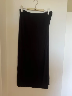 Svart långkjol med slits - En svart långkjol i mjukt material med en snygg slits på sidan. Kjolen har en enkel och stilren design som passar perfekt till många olika looks. Perfekt för dig som gillar minimalistisk stil och vill ha något bekvämt men ändå trendigt.