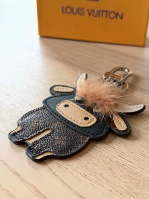 Vuitton nyckelring kofigur - Unik nyckelring från Vuitton i form av en söt kofigur. Tillverkad i brunt och beige läder med klassiskt LV-monogram på baksidan och fluffig beige detalj på huvudet. Perfekt accessoar till väskan eller nycklarna.