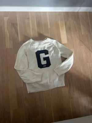Vit stickad tröja från Gant - Vit stickad tröja från Gant med stor mörkblå bokstav 'G' på bröstet. Tröjan har rund halsringning och ribbad nederkant. Perfekt för dig som gillar klassisk och sportig stil. Hör av dig till mig vid frågor 👍