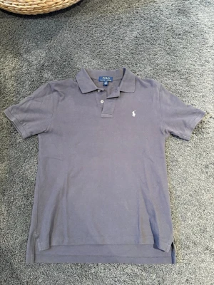 Mörkblå piké t-shirt - Klassisk mörkblå pikétröja från Polo Ralph Lauren med vit broderad logga på bröstet. Tröjan har korta ärmar, krage och knappar framtill. Perfekt till jeans eller shorts för en clean och stilren look. Riktigt jäkla snygg till absolut allting. Knappt använd men gammal så får lite vintage look. Tyvärr för liten för mig.