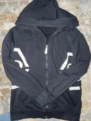 Hoodie Philipp plein - Säljer en svart hoodie med hel dragkedja och stor huva. På framsidan finns vita grafiska tryck i form av hexagoner med siffran 9 inuti på båda sidor. Hoodien har långa ärmar och ribbade muddar. Perfekt för en chill och stilren look.