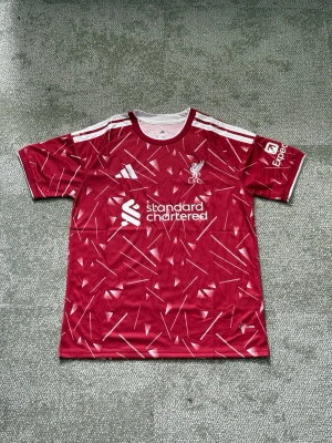 Liverpool röd matchtröja Adidas - Säljer en röd Liverpool matchtröja från Adidas med vitt geometriskt mönster, vita detaljer och klubbmärke på bröstet. Tröjan har korta ärmar, rund hals och är tillverkad i ett lätt, ventilerande material. Perfekt för fotboll eller träning.