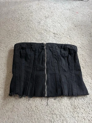 Svart denim bustierkorsett - Säljer en svart bustierkorsett i denim med dragkedja framtill och smockad rygg. Korsetten har råa, fransiga kanter nedtill för en edgy look och markerade sömmar som ger en snygg siluett. Perfekt att styla med jeans eller kjol.