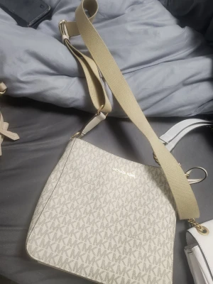 Beige axelväska från Michael Kors - Snygg beige axelväska från Michael Kors med justerbar bred axelrem och guldiga detaljer. Väskan har ett diskret MK-mönster över hela ytan och logotypen i guld framtill. Tillverkad i tåligt konstläder som ger en lyxig känsla.
