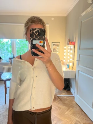 Vit kofta med pärlknappar - Söt vit kofta med kort ärm och pärlknappar framtill. Koftan har en avslappnad passform och ribbade kanter. Perfekt att slänga över ett linne eller klänning för en chill look. Har en knapp som saknas men finns en extra som går att sy dit 