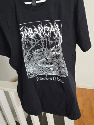 Cabancaya Hårdrockströja - Säljer en svart band-tshirt från Garbancya med tryck fram och bak. Framsidan har ett detaljerat vitt motiv och logga, baksidan har låtlista från albumet Presentiment Of Decay. Klassisk passform och rund hals. Perfekt för dig som gillar metal och unika bandtröjor.
