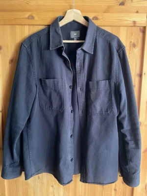 Svart overshirt från H&M - Stilren svart overshirt från H&M med klassisk krage och två stora bröstfickor. Skjortjackan har knäppning framtill och långa ärmar med knapp vid ärmslut. Tillverkad i ett kraftigare bomullstyg som ger en avslappnad look.