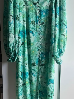Adoore klänning- mint green - Positano dress från Adoore använd en gång på grund av att jag tyvärr vuxit ur den. Långklänning i gröna och blå toner med blommigt mönster. Klänningen har långa puffärmar, knappar framtill och markerad midja i ett svalkande material. 