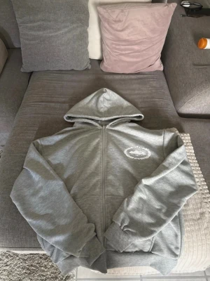 Grå zip hoodie från Cx - Snygg grå zip hoodie från Cx med huva och dragkedja framtill. Tröjan har ett tryckt vitt motiv på bröstet och ribbade muddar vid ärmar och nederkant. Perfekt för en avslappnad passform. Tillverkad i mjukt bomullsmaterial. Den är i storlek S men med perfekt passform för folk med M.
