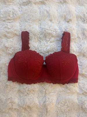 Röd Intimissimi bh med vaddering - Snygg röd Intimissimi bh med vadderade kupor och breda axelband i spets. Dekorativ spetsdetalj över hela kuporna och längs nederkanten. Bh:n har en liten rosett framtill, perfekt för dig som gillar feminina detaljer. Använd fåtal gånger. 