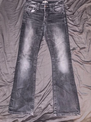 Grå bootcut jeans med slitningar - Säljer ett par grå bootcut jeans med snygga slitningar och tvättade detaljer. Jeansen har fem fickor, knappgylf och kontrastsömmar. Passar perfekt för dig som gillar en avslappnad men trendig stil.