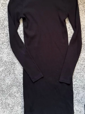Svart ribbad långärmad klänning - En enkel och stilren svart klänning med ribbad struktur och långa ärmar. Klänningen har en kroppsnära passform och är perfekt för dig som gillar minimalistisk stil. Passar till många olika tillfällen och är lätt att matcha med accessoarer.
