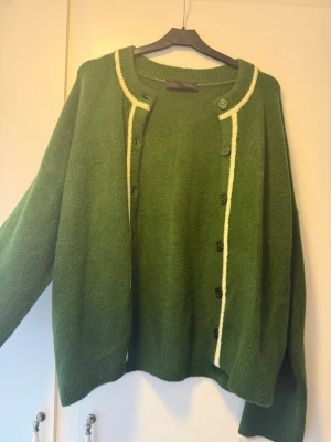 Grön kofta från ZARA med kontrastkant - Mysig grön kofta från ZARA med rund halsringning och knäppning framtill. Koftan har en vit kontrastkant längs knappraden och halsen, samt ribbade muddar vid ärmslut och nederkant. Perfekt för lager på lager och en chill look.