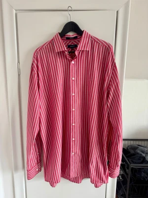 Rödvit randig skjorta från Gant - Säljer en röd och vit randig skjorta från Gant med klassisk krage och knappar framtill. Skjortan har lång ärm och är tillverkad i mjuk bomull. Broderad Gant-logga på bröstet för en clean look.