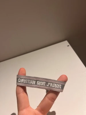 Grått Christian Dior J'Adior armband - Snyggt grått tygarmband från Christian Dior med texten 'J'Adior' broderad i vitt. Armbandet har justerbar knytning med tofsar i ändarna och en stilren, modern look som passar perfekt till din outfit.