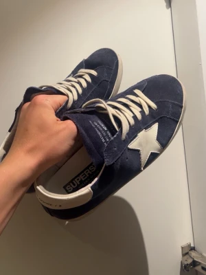 Mörkblå sneakers från Golden Goose - Snygga mörkblå sneakers från Golden Goose med vit stjärndetalj på sidan och vita skosnören. Ovandelen är i mocka och sulan är vit med svart yttersula. Klassisk låg modell med cool vintagekänsla och tydlig branding på insidan. Obs dom är i helt nytt skick🙌🙌