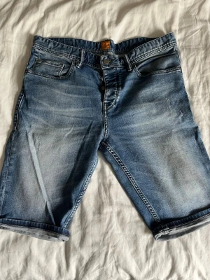 Blå jeansshorts från BOSS Orange - Snygga blå jeansshorts från BOSS Orange med klassisk femficksmodell och uppvikta benslut. Shortsen har en avslappnad passform och är tillverkade i slitstark bomull med snygga slitningar och tvättade detaljer. Perfekta för sommardagar och chill häng.
