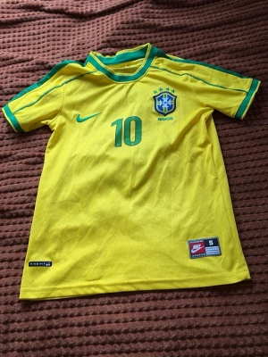 Brasilien fotbollströja Ronaldinho - Gul Brasilien fotbollströja från Nike med gröna detaljer, nummer 10 och Ronaldinho på ryggen. Tröjan har korta ärmar, rund hals och officiellt landslagsmärke på bröstet. Perfekt för dig som älskar fotboll och vill bära en ikonisk matchtröja.