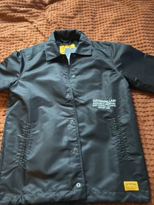 Svart vindjacka från Caterpillar - Svart vindjacka från Caterpillar med tryck på bröstet och gul logotyp vid nederkanten. Nypris 1600. Jackan har klassisk krage, knäppning med tryckknappar och två sidofickor. Materialet är glansigt och vattenavvisande, perfekt för blåsiga dagar.