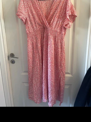 Rosa blommig långklänning  - Söt rosa långklänning med små vita blommor och omlott-skärning. Klänningen har korta ärmar, v-ringning och smock i midjan för en bekväm passform. Perfekt för varma dagar och somriga tillfällen.