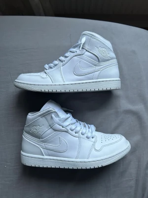 Vita Nike Air Jordan 1 Mid sneakers - Säljer ett par helvita Nike Air Jordan 1 Mid sneakers i skinn med klassisk swoosh på sidan och Air Jordan-logga vid ankeln. Skorna har vita skosnören, perforerad tåbox och en stabil sula. Perfekta för dig som gillar stilrena och ikoniska sneakers.