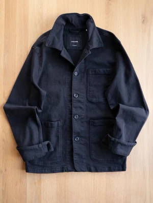 Svart overshirt med stora fickor - Stilren svart overshirt i kraftigt bomullstyg med tre stora utanpåfickor och knäppning framtill. Klassisk krage och avslappnad passform, perfekt att slänga på sig över en t-shirt. Snygg och enkel design som funkar till det mesta.