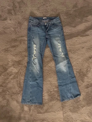 Blå bootcut jeans med slitningar och rosa detaljer - Säljer ett par ljusblå bootcut jeans från Gina Young i storlek 170,  de har slitningar framtill och broderade detaljer på bakfickorna. Jeansen har klassisk femficksmodell och är tillverkade i jeansmaterial. Perfekta för dig som gillar en avslappnad och trendig stil.