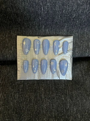 Press-on naglar || Storlek L - Snygga press-on naglar i en himmelblå, glittrig färg med silverfärgade stjärnor, månar och linjer som motiv. Naglarna har en mandelformad design och är perfekta för dig som vill ha en unik och trendig look. Storlek L, Det du får med dig : * Fil och manikyr pinne * nagellim klistermärken * 2 lappar av rengöring för naglar.