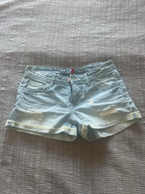 Ljusblå denimshorts med slitningar - Säljer ett par ljusblå denimshorts med slitna detaljer och uppvikta benslut. Klassisk femficksmodell med knapp och dragkedja framtill. Perfekta för varma dagar och ger en avslappnad look.
