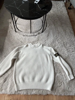 Vit stickad tröja från Stone Island - Säljer en stilren vit stickad tröja från Stone Island med ribbad struktur och rund hals. Tröjan har lång ärm och den klassiska Stone Island-loggan på vänster ärm. Perfekt för dig som gillar minimalistisk och clean stil.