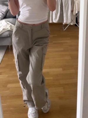 Beiga cargobyxor med fickor - Säljer ett par beiga cargobyxor med raka ben och stora sidofickor. Byxorna har en avslappnad passform och stängs med knapp och dragkedja. Perfekta för en casual och trendig look. Materialet ser ut att vara bomull eller bomullsblandning.