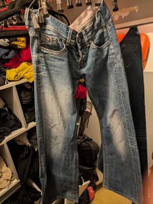 Blå bootcut jeans med slitningar från replay - Säljer ett par blå bootcut jeans med snygga slitningar och ljusare partier framtill. Klassisk femficksmodell med knappgylf och kontrastsömmar. Jeansen har en avslappnad passform och är perfekta för en casual look. Materialet är denim i bomull.