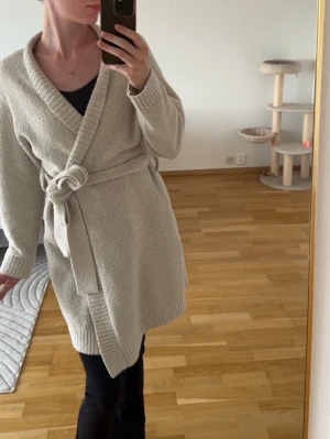 Beige omlott kofta med bälte - Mysig beige kofta i omlottmodell med knytskärp i midjan. Tillverkad i ett mjukt, stickat material som känns skönt mot huden. Perfekt för lata morgnar eller kvällar hemma. Lång ärm och ribbade muddar för extra komfort.