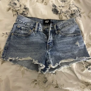 Blå jeansshorts från 157, XXS - Säljer ett par blå jeansshorts från 157 i storlek XXS men passar även folk med XS. De har råa, fransiga kanter och slitna detaljer framtill för en avslappnad look. Klassisk femficksmodell med knapp och dragkedja. Perfekta till sommaren!