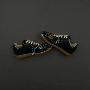 Svarta Maison Margiellas med färgstänk - Unika svarta sneakers från Maison Margiela med färgstänk. Skorna har vita snören, beige innersula och klassisk gummisula. Låg modell med cool konstnärlig vibe och retroinspirerad siluett. Perfekta för dig som vill sticka ut.