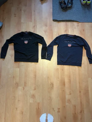Två sweatshirts från GANT - Två  sweatshirts från GANT en marin blå och en svart med broderad logga och sköld på bröstet. Klassisk rund halsringning och långa ärmar. Tröjorna har ribbade muddar vid ärmslut och nederkant. Perfekt för en stilren och avslappnad look.